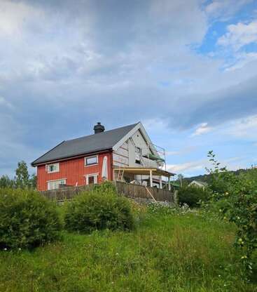 L'image représente une maison à deux étages avec un extérieur rouge et un toit gris, située sur une colline au milieu d'une végétation luxuriante et d'un ciel nuageux.