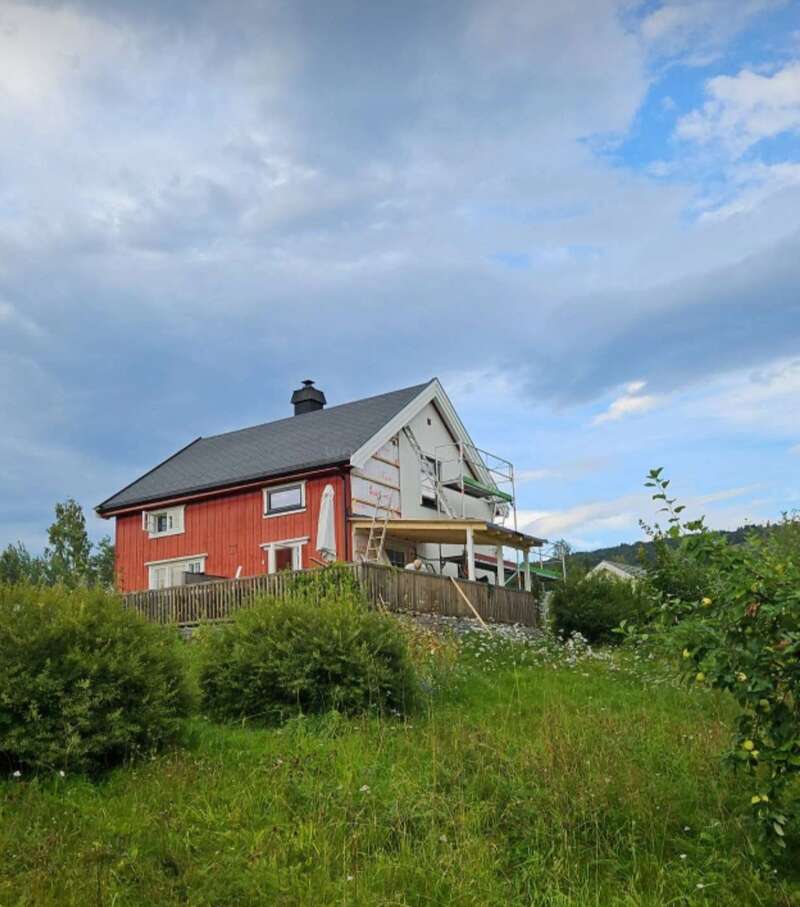 Das Bild zeigt ein zweistöckiges Haus mit roter Fassade und grauem Dach, das auf einem Hügel inmitten von üppigem Grün und einem bewölkten Himmel steht.