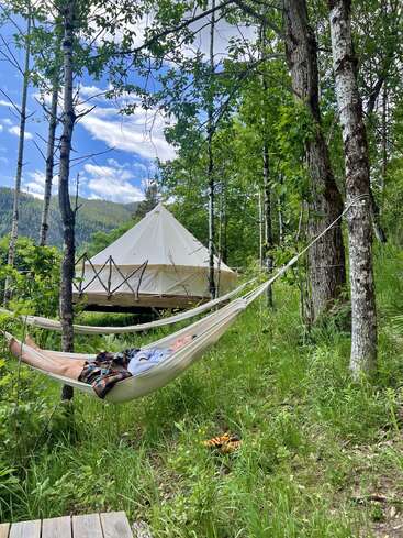 Une personne se détend dans un hamac au milieu de grands arbres et d'une végétation luxuriante, avec une tente confortable en arrière-plan. Le ciel est bleu avec des nuages épars.