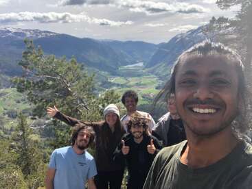 Un groupe d'amis sourit et pose pour un selfie lors d'une randonnée en montagne, avec en arrière-plan des vallées verdoyantes, des rivières et des montagnes sous un ciel nuageux.