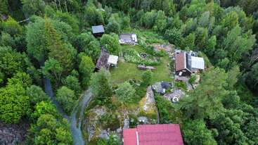 Cette image aérienne montre une scène de campagne paisible avec plusieurs bâtiments nichés au milieu d'arbres denses et verdoyants, entourés par la nature, des chemins sinueux et des jardins.