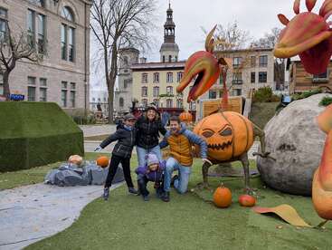 Una familia posa felizmente al aire libre junto a grandes y caprichosos adornos de Halloween, entre ellos una linterna y criaturas vegetales gigantes, con edificios históricos al fondo.