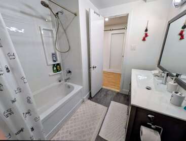 Este cuarto de baño tiene una bañera blanca, una bonita cortina de ducha con motivos de perros, un tocador con espejo, dos lavabos, alfombrillas y algunos adornos colgantes rojos en la pared.