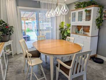 Un acogedor comedor con una mesa de madera, cuatro sillas, plantas, modernas lámparas colgantes, armarios blancos, puertas de cristal y asientos al aire libre visibles a través de puertas correderas.