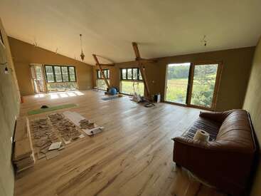 A spacious, sunlit room with wooden floors, large windows, and exposed beams. A brown sofa, yoga mat, exercise equipment, and scattered papers furnish the space.