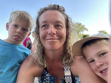 The image shows a woman posing for a selfie with two young boys, set against a clear blue sky with trees in the background.