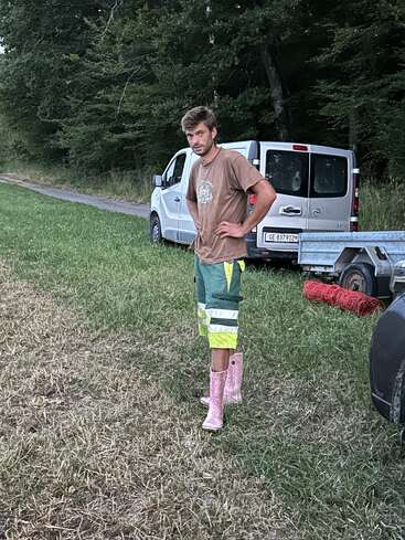 The image depicts a man standing in a grassy field, wearing a brown t-shirt, shorts, and pink boots, with a white van and trailer in the background.
