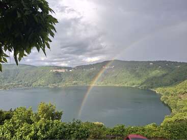 A serene lake surrounded by lush green hills, under a cloudy sky, features a vibrant rainbow arching above the water, framed by leafy branches. Peaceful scene.