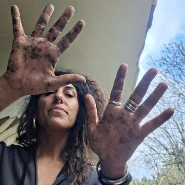 A woman with curly hair and rings shows her dirty hands to the camera, likely from gardening or outdoor work. The background shows a clear sky.