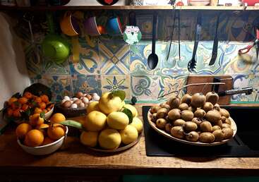 Una acogedora escena de cocina con cuencos de naranjas frescas, limones, kiwis y huevos sobre una encimera de madera, bajo tazas de colores y un salpicadero de azulejos estampados.