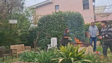 Tres personas disfrutan de una barbacoa en un frondoso jardín con arbustos verdes, bancos de madera y una casa rosa al fondo en un día nublado.
