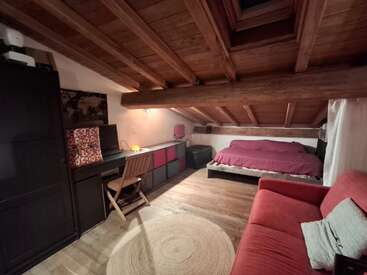 Esta acogedora habitación abuhardillada cuenta con un techo inclinado de madera, una cama doble con ropa de cama roja, un escritorio, un sofá rojo, armarios, una manta mullida y una iluminación cálida.