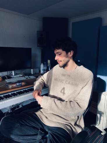 A young man sits in a cozy music studio, wearing a beige sweater. He is relaxed, surrounded by monitors, keyboards, and other music production equipment.