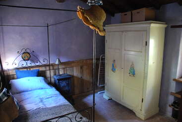 This cozy bedroom features a single iron bed, a vintage white wardrobe with rabbit paintings, a small stove, purple walls, and a hanging straw hat.