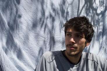 Un joven de pelo oscuro y barba corta se sienta pensativo delante de un fondo de tela a rayas, con la luz del sol moteada y las sombras creando un efecto artístico.