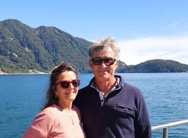 A smiling couple wearing sunglasses stands together by a scenic lake with clear blue water, lush green hills, and a bright, sunny sky in the background.