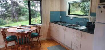 This cozy kitchen features white cabinets, a small round dining table with four chairs, large windows with green views, a gas stove, sink, and refrigerator.