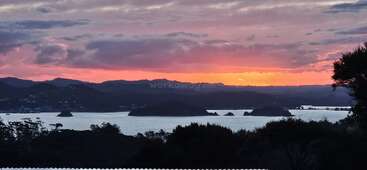 Dieses Bild zeigt einen farbenfrohen Sonnenuntergang über einer ruhigen Bucht mit verstreuten Inseln, silhouettierten Hügeln und dramatischen Wolken.