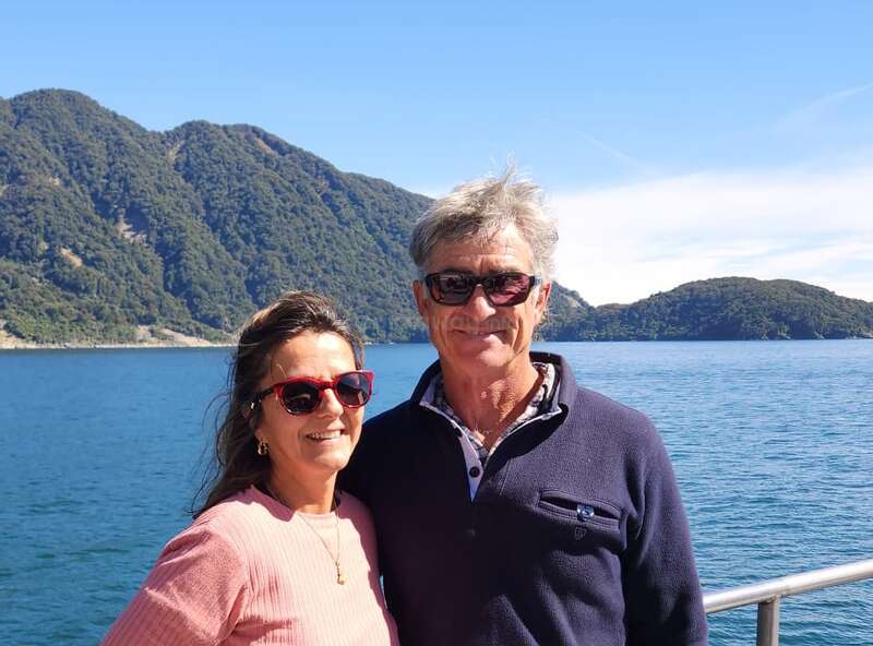 A smiling couple wearing sunglasses stands together by a scenic lake with clear blue water, lush green hills, and a bright, sunny sky in the background.
