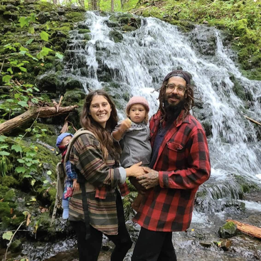 The image depicts a family of three standing in front of a waterfall, with the mother carrying a baby on her back and the father holding their other child.