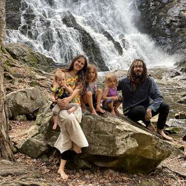 Una feliz familia de cinco miembros, descalza, está sentada sobre unas rocas frente a una preciosa cascada en plena naturaleza. Todos sonríen y disfrutan juntos del entorno al aire libre.