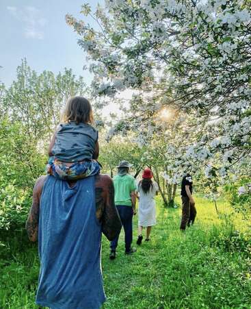 Eine Gruppe spaziert durch einen sonnigen, blühenden Obstgarten. Ein Kind sitzt auf den Schultern eines Erwachsenen, umgeben von üppigem Grün und blühenden weißen Blumen, und genießt gemeinsam die Natur.