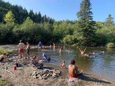 Eine Gruppe von Leuten genießt es, an einem sonnigen Tag unter strahlend blauem Himmel in einem seichten Fluss zu schwimmen und sich zu entspannen, umgeben von üppig grünen Bäumen.