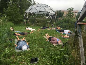 Eine Gruppe von Menschen liegt draußen auf Matten im Gras, in der Nähe einer geodätischen Kuppel, umgeben von viel Grün, und scheint an einer Entspannungs- oder Meditationssitzung teilzunehmen.