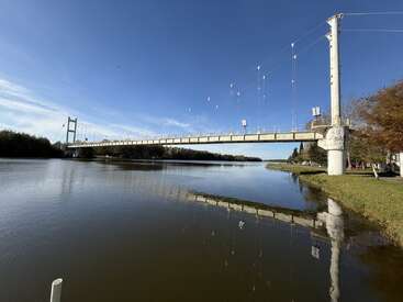 Eine Hängebrücke überspannt das ruhige Wasser des Flusses und spiegelt seine Struktur wider. Klarer blauer Himmel, grüne Bäume und Gras säumen die Flussufer an einem sonnigen Tag.