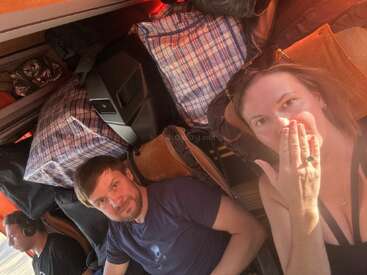 Drei Personen sitzen zusammen in einem überfüllten Fahrzeug, umgeben von großen karierten Taschen. Eine Frau schaut überrascht und verdeckt ihren Mund, während zwei Männer nach vorne schauen.