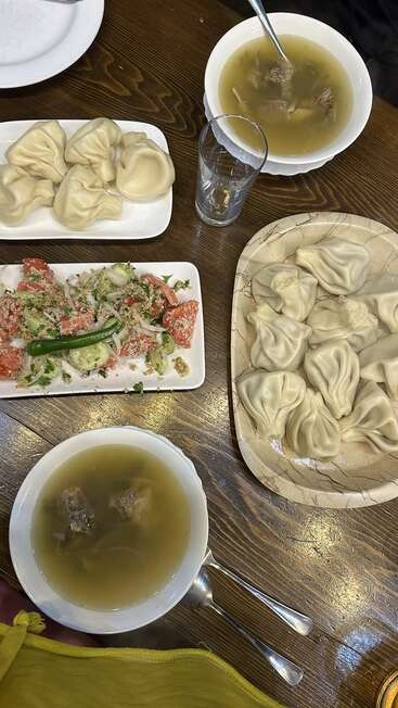 Dieses Bild zeigt einen gedeckten Holztisch mit Suppenschüsseln, Tellern mit Khinkali-Klößen, einem frischen Salat, Besteck und einem klaren Glas Wasser.