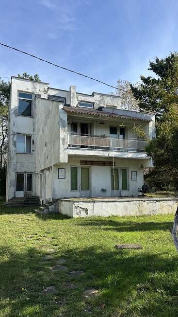 Ein zweistöckiges, verwittertes weißes Gebäude mit abblätternder Farbe und sichtbaren Rissen. Umgeben von Gras, Bäumen und einem blauen Himmel wirkt es etwas vernachlässigt und verlassen.