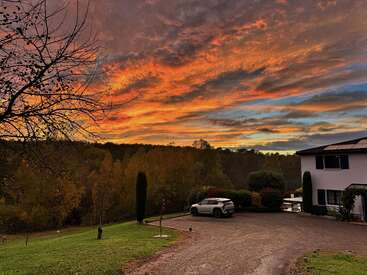 Una impresionante puesta de sol llena el cielo de vibrantes tonos naranjas y morados. Un moderno coche blanco está aparcado junto a una casa blanca, rodeada de árboles y vegetación.