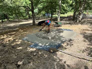 Uma pessoa está medindo concreto recém-despejado sobre uma lona em uma área ao ar livre, sombreada e arborizada, com ferramentas e um carrinho de mão por perto, sob a luz forte do dia.