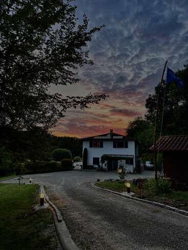 Um lindo céu ao pôr do sol, repleto de nuvens coloridas, domina o cenário. Uma aconchegante casa branca ergue-se no final de uma estrada sinuosa, cercada por vegetação.