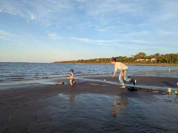 Ein Kind und ein Erwachsener spielen und rennen an einem Sandstrand in der Nähe des Wassers, mit klarem Himmel, sanften Wellen und fernen Bäumen im Hintergrund.