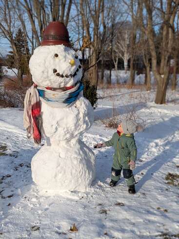 Ein kleines Kind in Winterkleidung bewundert einen großen Schneemann mit Schal, Blumentopfmütze und lächelndem Gesicht in einem verschneiten, sonnenbeschienenen Park, der von Bäumen umgeben ist.
