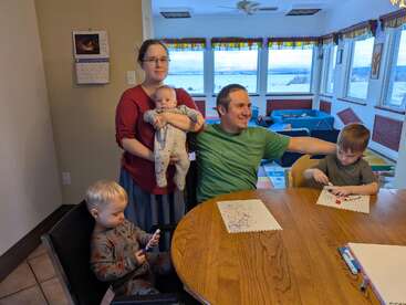 Eine fünfköpfige Familie versammelt sich um einen Holztisch. Zwei kleine Kinder malen mit Stiften, während ein Baby im Arm gehalten wird. Große Fenster geben den Blick auf eine verschneite Landschaft frei.