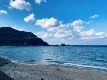 Una playa tranquila con suaves olas se encuentra con una orilla de guijarros, exuberantes colinas verdes se elevan en el fondo, y esponjosas nubes blancas flotan a través de un cielo azul brillante.