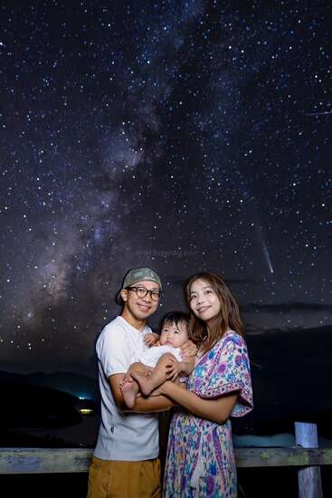 Una sonriente familia de tres miembros bajo un impresionante cielo estrellado. Los padres sostienen cariñosamente a su bebé sobre un fondo de estrellas parpadeantes y la Vía Láctea.