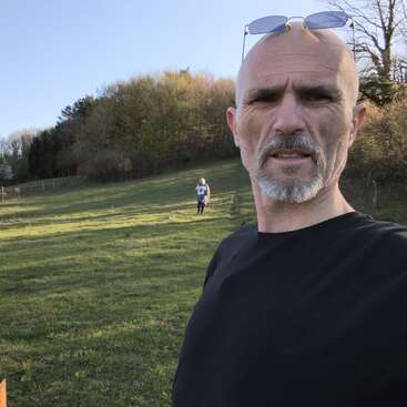 A man with sunglasses on his head takes a selfie in a grassy field with trees. Another person is walking in the background under clear skies.