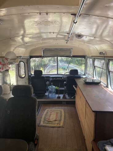The image shows the inside of a converted school bus with cozy seating, a rug, wooden cabinets, water jug, kitchen supplies, and natural light streaming in.