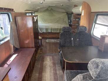 This image shows the interior of a converted bus with wood paneling, carpet runners, a small table with chairs, cabinets, and a bed with a mosquito net.