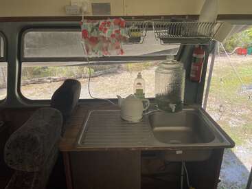 This image shows a compact kitchen inside a vehicle. There’s a sink, kettle, soap, glass water dispenser, drying rack, and a window with seats nearby.