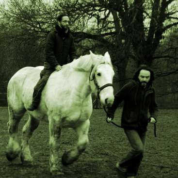 L'image représente deux hommes interagissant avec un cheval blanc dans un cadre extérieur, l'un montant le cheval et l'autre le menant par les rênes.