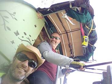 L'image montre deux hommes posant pour un selfie devant une tente verte avec un chapeau jaune et un homme en chemise rouge tenant un arc et des flèches.
