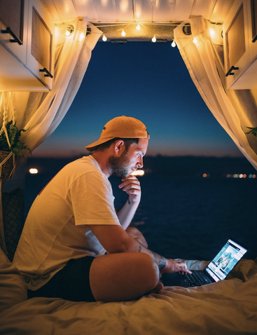 Ein Mann sitzt auf einem Bett in einem Wohnmobil und schaut auf einen Laptop, während er durch offene Vorhänge den Nachthimmel sieht.