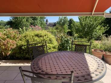 A sunny patio with a patterned round table and chairs sits under a red awning, facing lush green bushes, trees, potted plants, and distant rooftops.