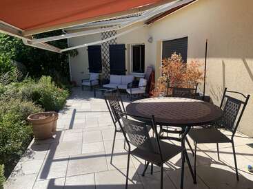 A sunny patio features a round table with four chairs, potted plants, a seating area with a loveseat, an awning for shade, and lush surrounding greenery.