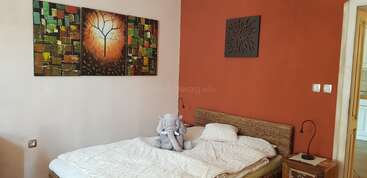 Ein gemütliches Schlafzimmer mit einem Doppelbett, weißer Bettwäsche und einem Plüschelefanten. Bunte abstrakte Kunst und eine rote Akzentwand schaffen eine warme, einladende Atmosphäre.